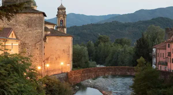 Ponte in pietra sul fiume, con edifici storici illuminati e il monte Argenia sullo sfondo.