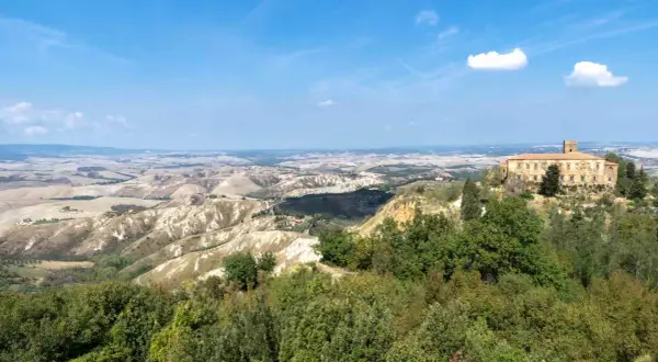 Panoramica delle balze di Volterra, con un antico palazzo in pietra su una collina e dolci colline ondulate all'orizzonte sotto un cielo azzurro.