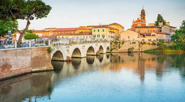 Il Ponte di Tiberio a Rimini, con i suoi archi in pietra che si specchiano sull'acqua calma, fa da sfondo a un paesaggio urbano pittoresco.