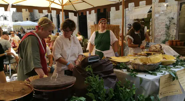 Donne in costume d'epoca servono cibo a un **Volterra markt**. Cesti, pentole e verdure fresche decorano il banco.