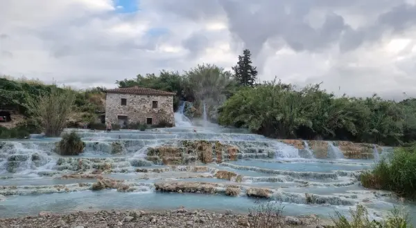 Cascate naturali in Toscana con un antico casale in pietra. L'acqua azzurra scorre su rocce calcaree, creando piscine termali dove alcune persone si rilassano.