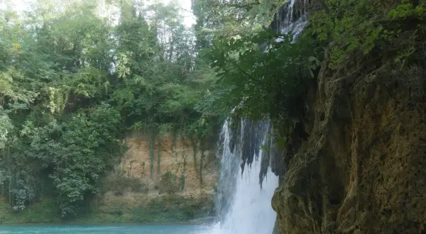 Cascata nel parco fluviale dell'Elsa, con acqua turchese e vegetazione lussureggiante che incornicia la roccia.