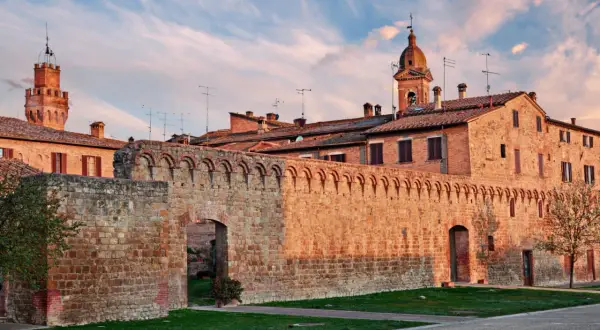 Antica cinta muraria e torri in mattoni rossi sotto un cielo azzurro striato di nuvole rosa, tipico delle colline senesi al tramonto.