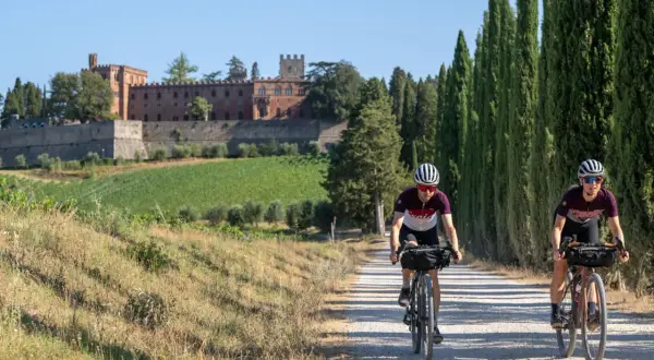 Due ciclisti affrontano un eroico percorso su una strada sterrata in Toscana, con un castello e vigneti sullo sfondo.