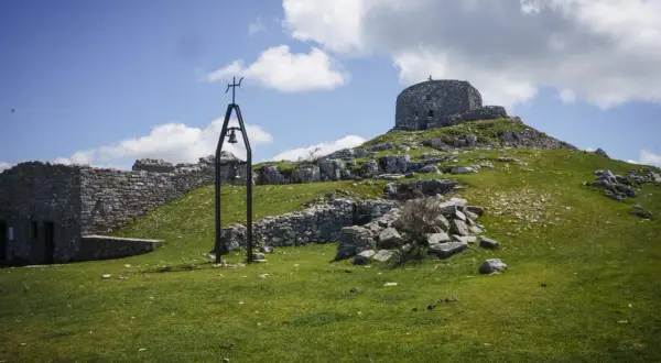Rovine di pietra e una campana sul **monte Labbro**, sotto un cielo azzurro con nuvole bianche.