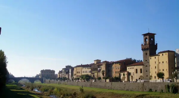 Pescia: cosa vedere? Un fiume serpeggia tra edifici storici e una torre imponente, sotto un cielo azzurro.