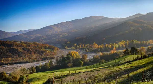 Colline autunnali illuminate dal sole, perfette per il trekking in Emilia Romagna. Un fiume serpeggia tra alberi dai colori caldi.