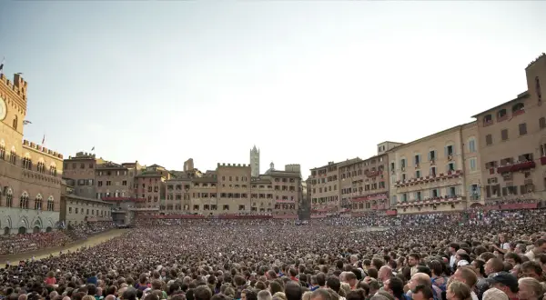 Folla immensa in Piazza del Campo a Siena, in attesa di sapere quand'è il Palio. Edifici storici e cielo terso.