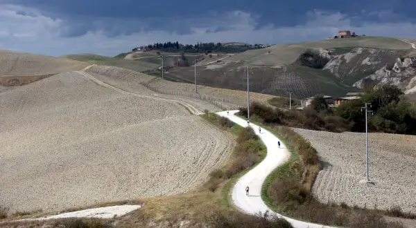 Ciclisti affrontano una strada eroica toscana, serpeggiando tra colline arate sotto un cielo minaccioso.