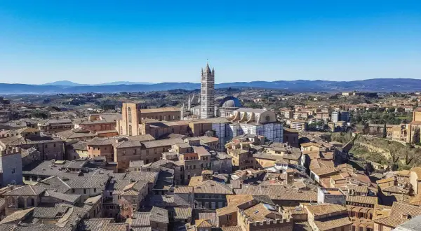 Panoramica di **Siena da vedere**: tetti in cotto, il Duomo e il Campanile svettano su un paesaggio collinare sotto un cielo azzurro.