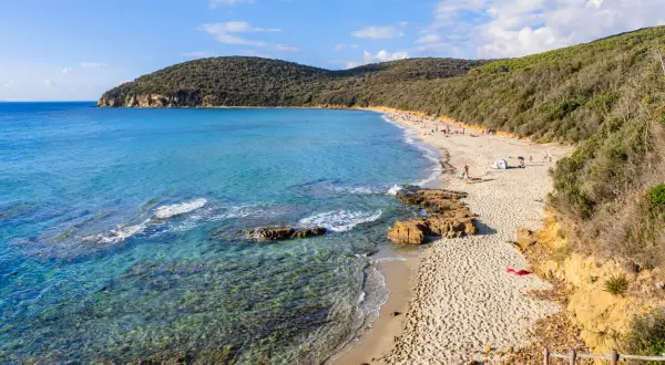 Spiaggia delle Bandite di Scarlino con acque cristalline, sabbia dorata e vegetazione rigogliosa. Persone rilassate al sole.