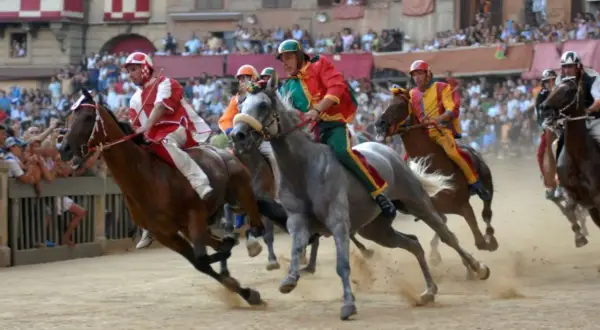 Cavalli e fantini in gara durante il Palio di Siena. La polvere si solleva mentre i cavalli corrono verso la vittoria.