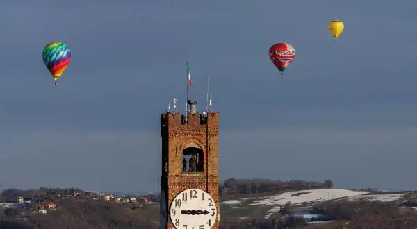 Un colorato evento mongolfiere vola nel cielo azzurro sopra una torre dell'orologio e un paesaggio collinare innevato.