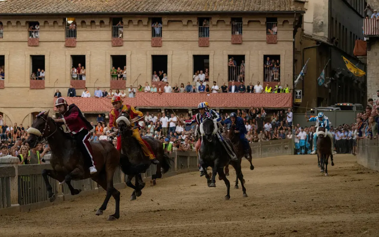 Cavalli e fantini in gara durante le contrade di Siena, con spettatori affacciati ai palazzi.