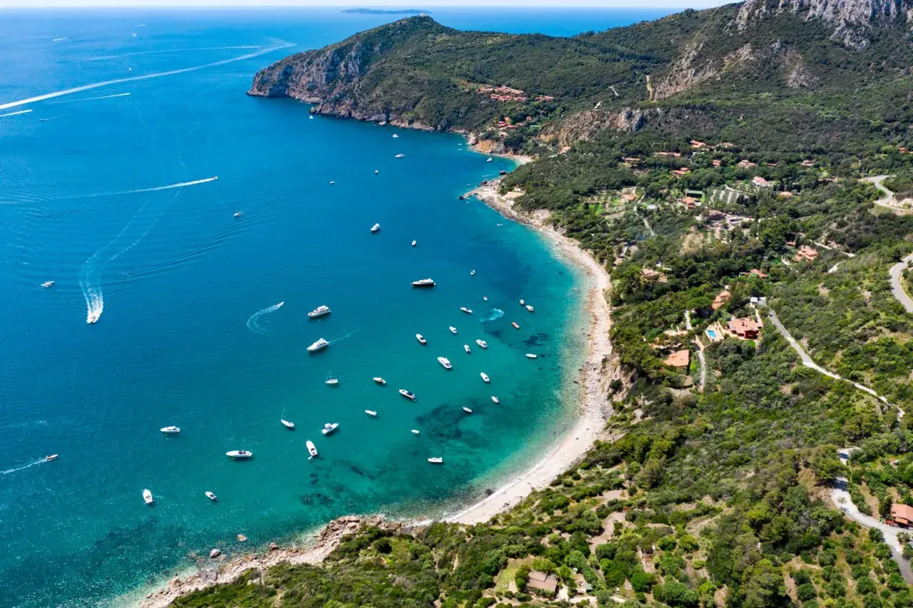Baia con acque cristalline e numerose barche al largo della costa del Monte Argentario. Ville immerse nel verde e una strada panoramica.