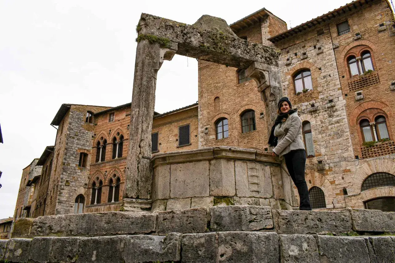 Donna sorride vicino a un antico pozzo a San Gimignano, con palazzi medievali sullo sfondo.