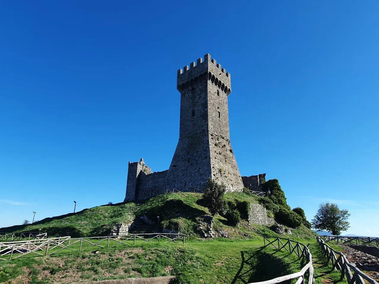 La fortezza di Radicofani domina il paesaggio sotto un cielo azzurro. Le antiche mura e la torre si ergono su una collina verdeggiante.