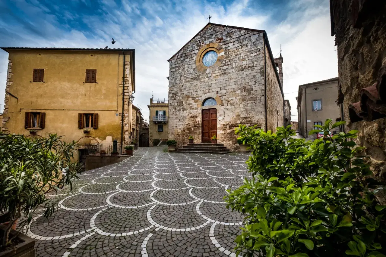 Piazza con pavimentazione a ventaglio, chiesa in pietra e case gialle, tipici borghi vicino Marina di Bibbona.