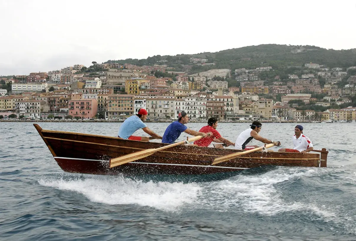 Equipaggio in gara nel palio marinaro argentario, vogando all'unisono con la città sullo sfondo.