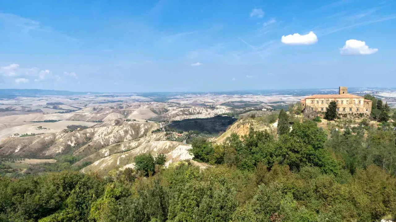 Panoramica delle balze di Volterra, con un antico palazzo in pietra su una collina e dolci colline ondulate all'orizzonte sotto un cielo azzurro.