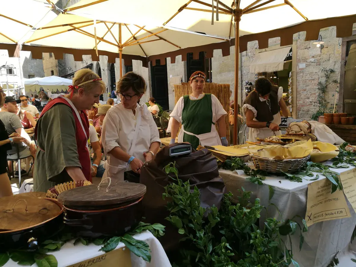 Donne in costume d'epoca servono cibo a un **Volterra markt**. Cesti, pentole e verdure fresche decorano il banco.