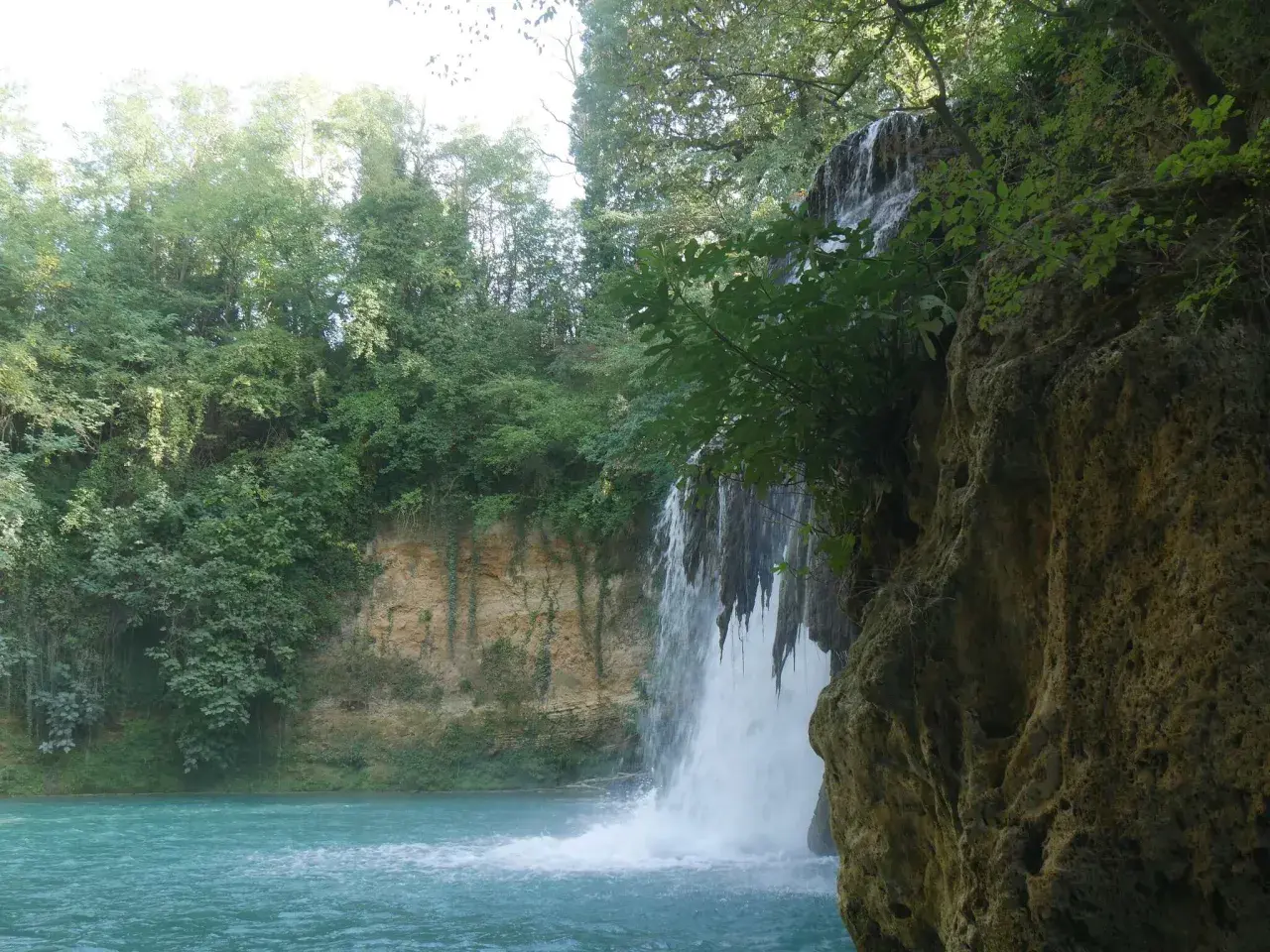 Cascata nel parco fluviale dell'Elsa, con acqua turchese e vegetazione lussureggiante che incornicia la roccia.