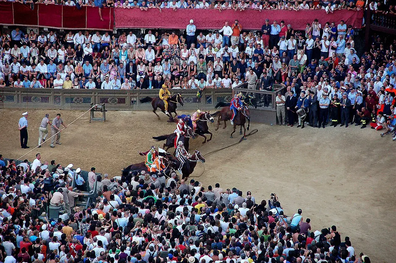 Cavalli e fantini in gara durante il Palio dell'Assunta a Siena, tra la folla festante.