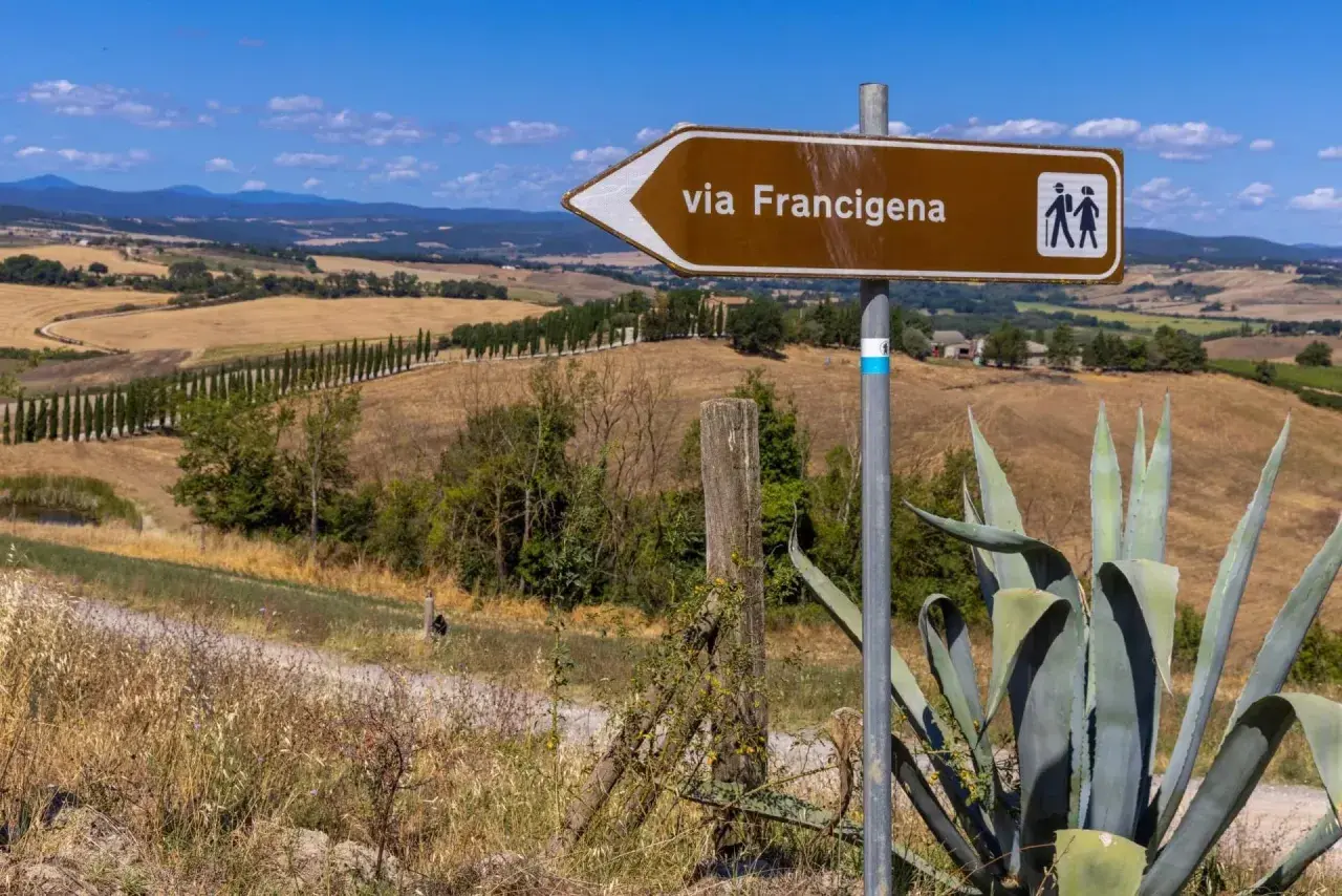 Segnale marrone indica la via Francigena, con un paesaggio collinare toscano sullo sfondo.