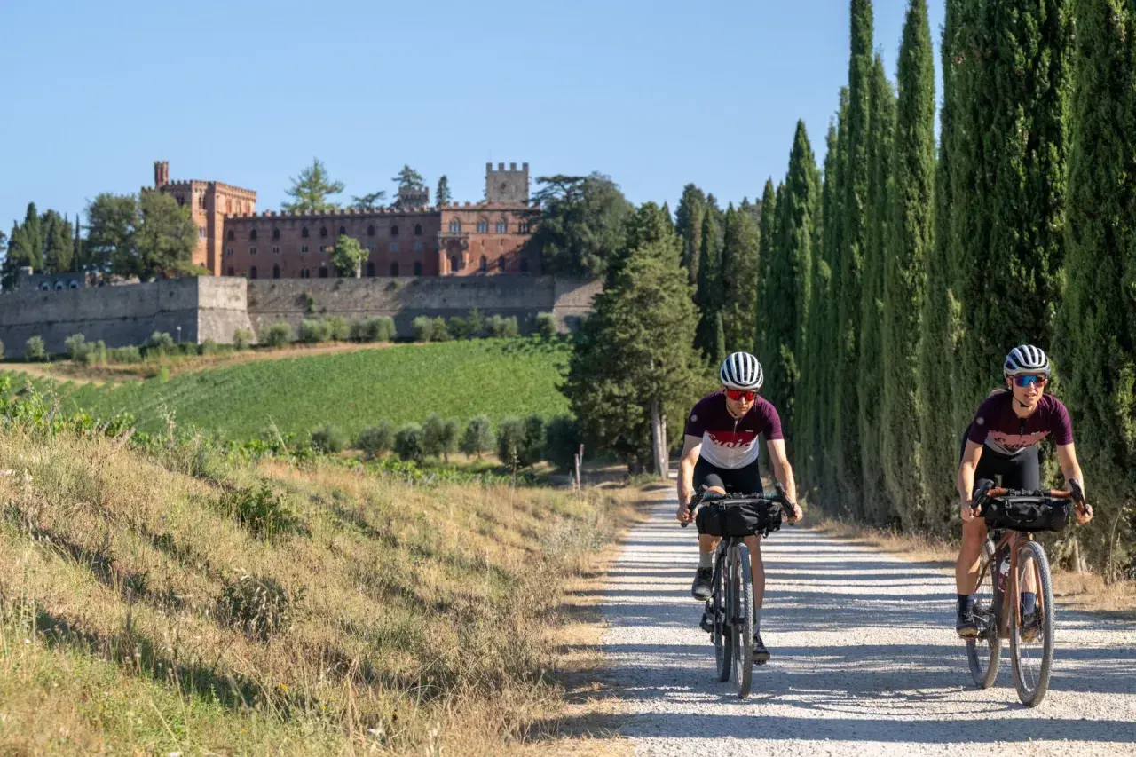 Due ciclisti affrontano un eroico percorso su una strada sterrata in Toscana, con un castello e vigneti sullo sfondo.