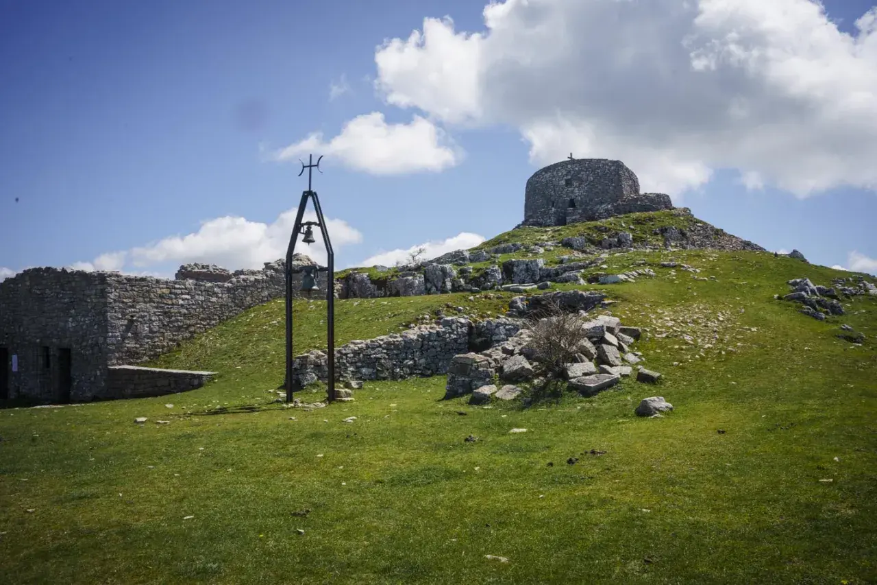 Rovine di pietra e una campana sul **monte Labbro**, sotto un cielo azzurro con nuvole bianche.