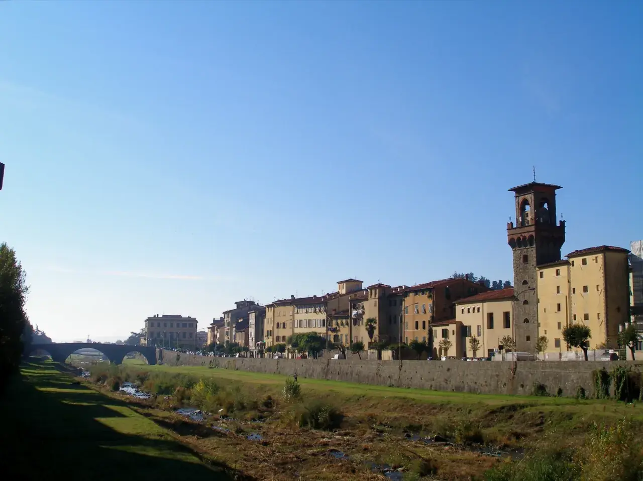 Pescia: cosa vedere? Un fiume serpeggia tra edifici storici e una torre imponente, sotto un cielo azzurro.