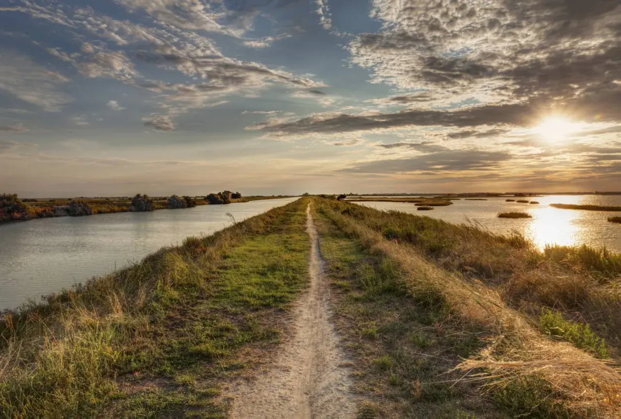 Sentiero erboso lungo il percorso del Po, con acque tranquille e cielo nuvoloso al tramonto.