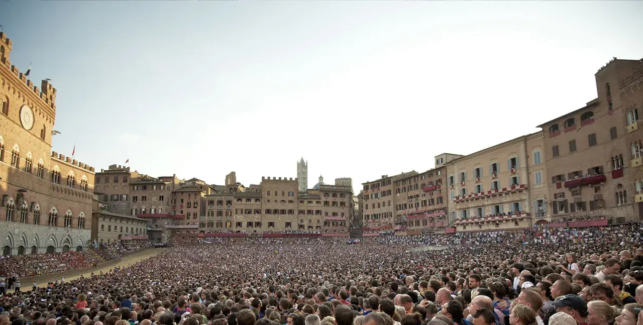 Folla immensa in Piazza del Campo a Siena, in attesa di sapere quand'è il Palio. Edifici storici e cielo terso.