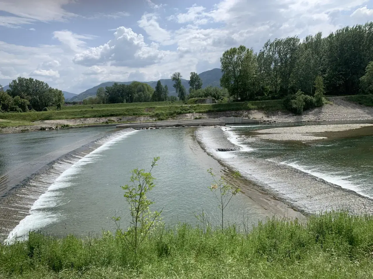 Il **parco fluviale del Serchio** offre uno spettacolo di acqua che scorre su dighe naturali, circondato da verdi colline e alberi rigogliosi sotto un cielo nuvoloso.