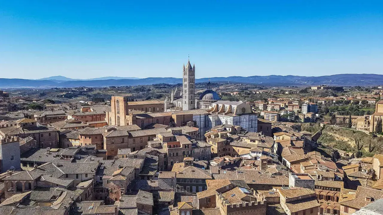 Panoramica di **Siena da vedere**: tetti in cotto, il Duomo e il Campanile svettano su un paesaggio collinare sotto un cielo azzurro.