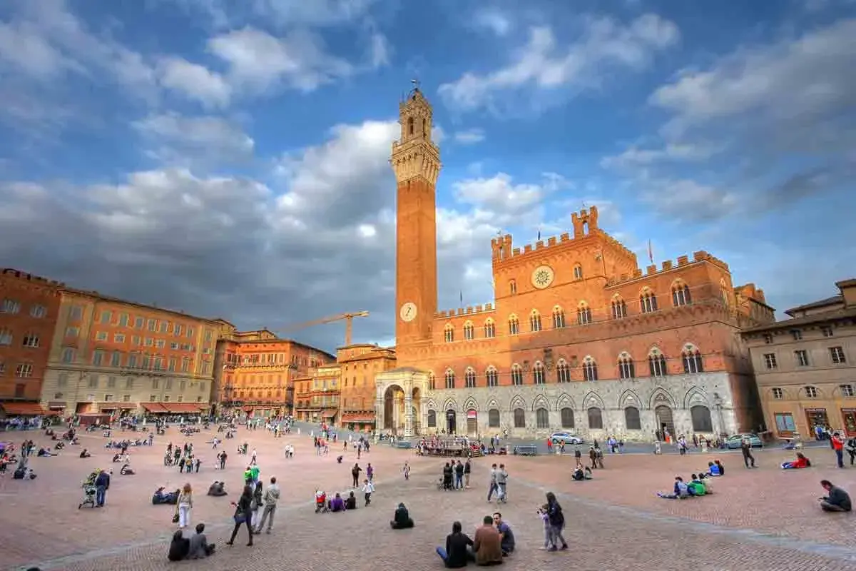 Piazza del Campo a Siena, un luogo iconico per il turismo, con il Palazzo Pubblico e la Torre del Mangia sotto un cielo nuvoloso.