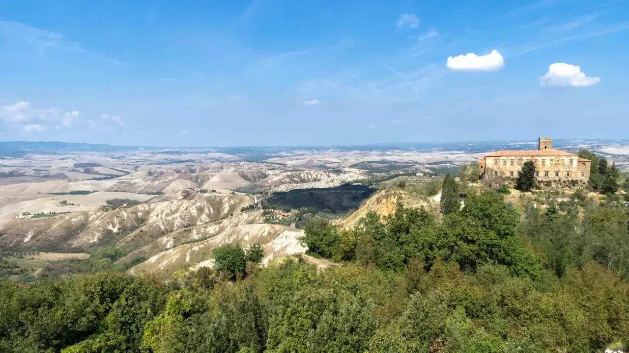 Panoramica delle balze di Volterra, con un casale in pietra in primo piano e colline ondulate all'orizzonte sotto un cielo azzurro.