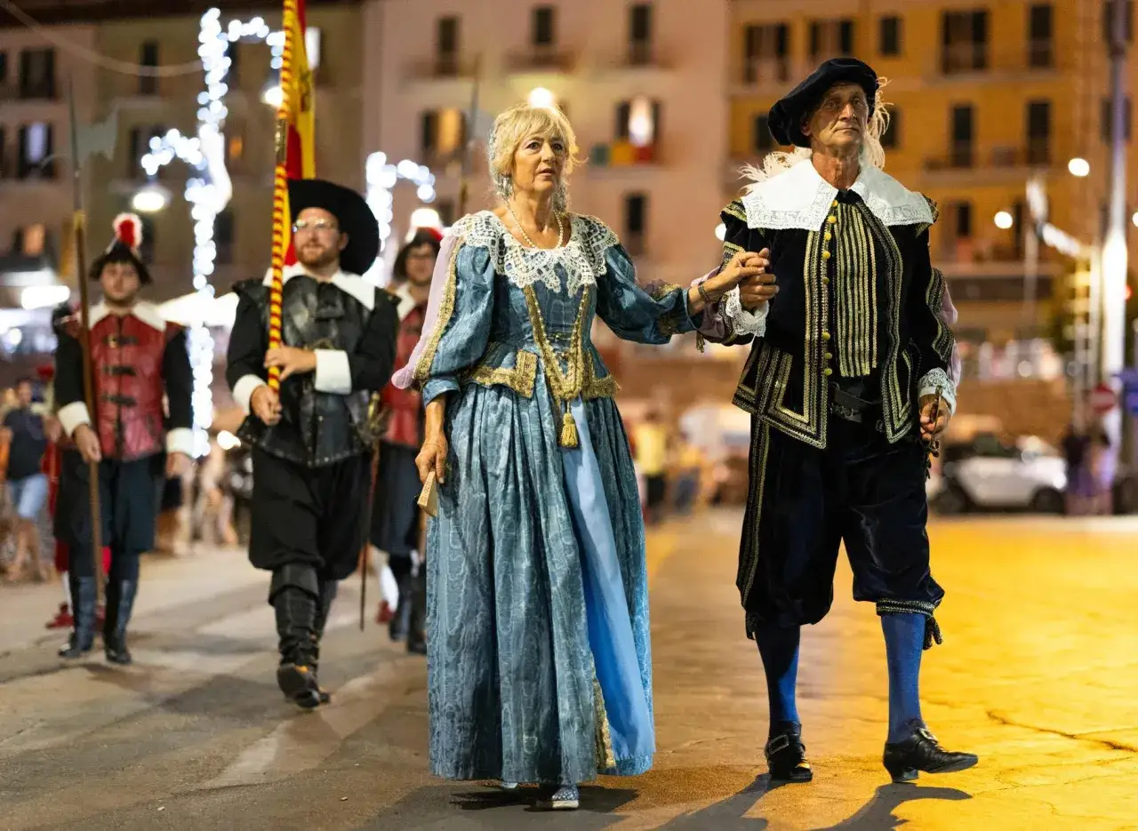 Coppia in costume storico sfila durante il Palio Marinaro Argentario. Uomini in divisa d'epoca li seguono.