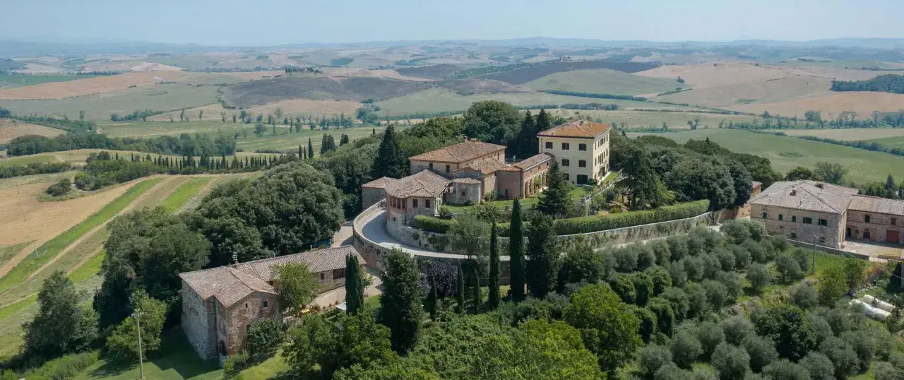 Un complesso di edifici in pietra e terracotta, immerso nel verde, ricorda i borghi vicino Parma, con dolci colline sullo sfondo.