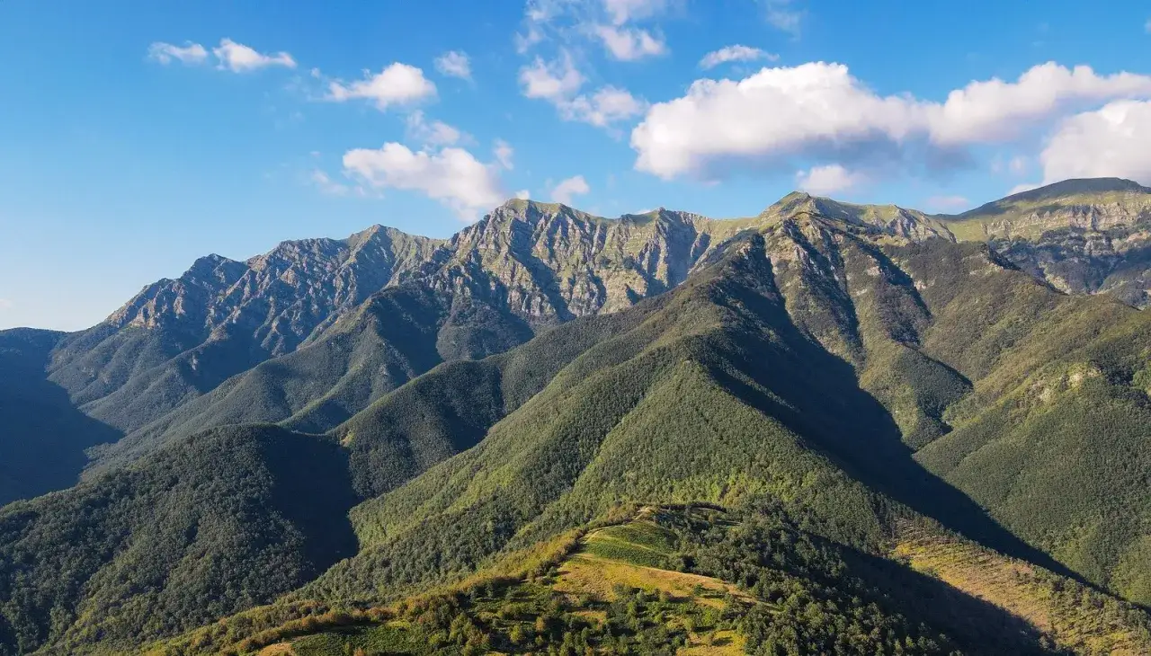 Vaste montagne dell'Emilia Romagna, con cime rocciose e pendii boscosi sotto un cielo azzurro con nuvole bianche.