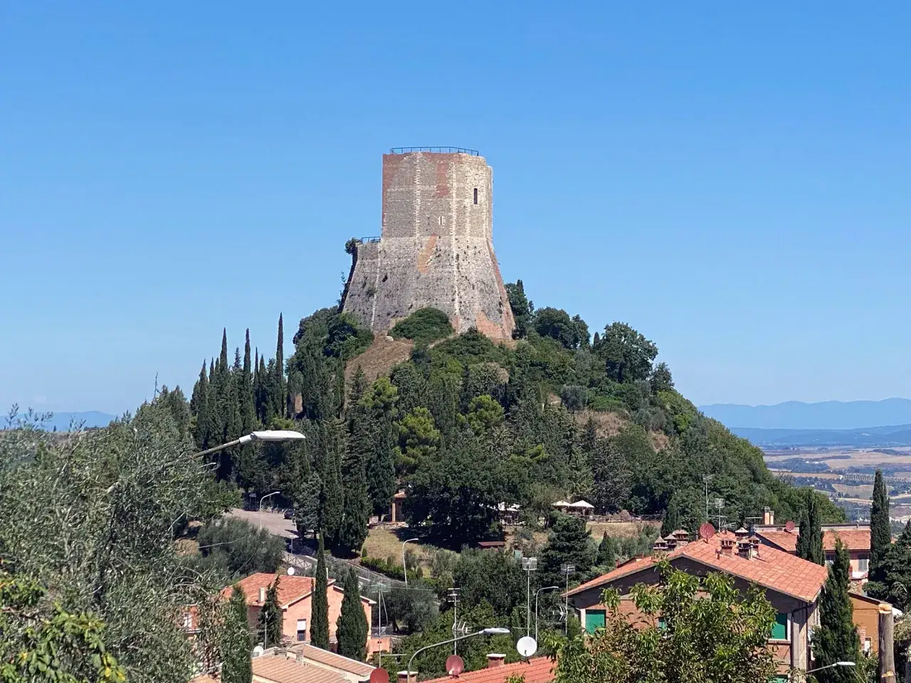 La rocca di Tentennano domina la collina, circondata da cipressi e case con tetti rossi, sotto un cielo azzurro.