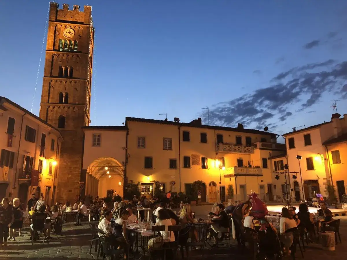 Serata ad Altopascio, con la torre illuminata e gente seduta ai tavoli all'aperto.