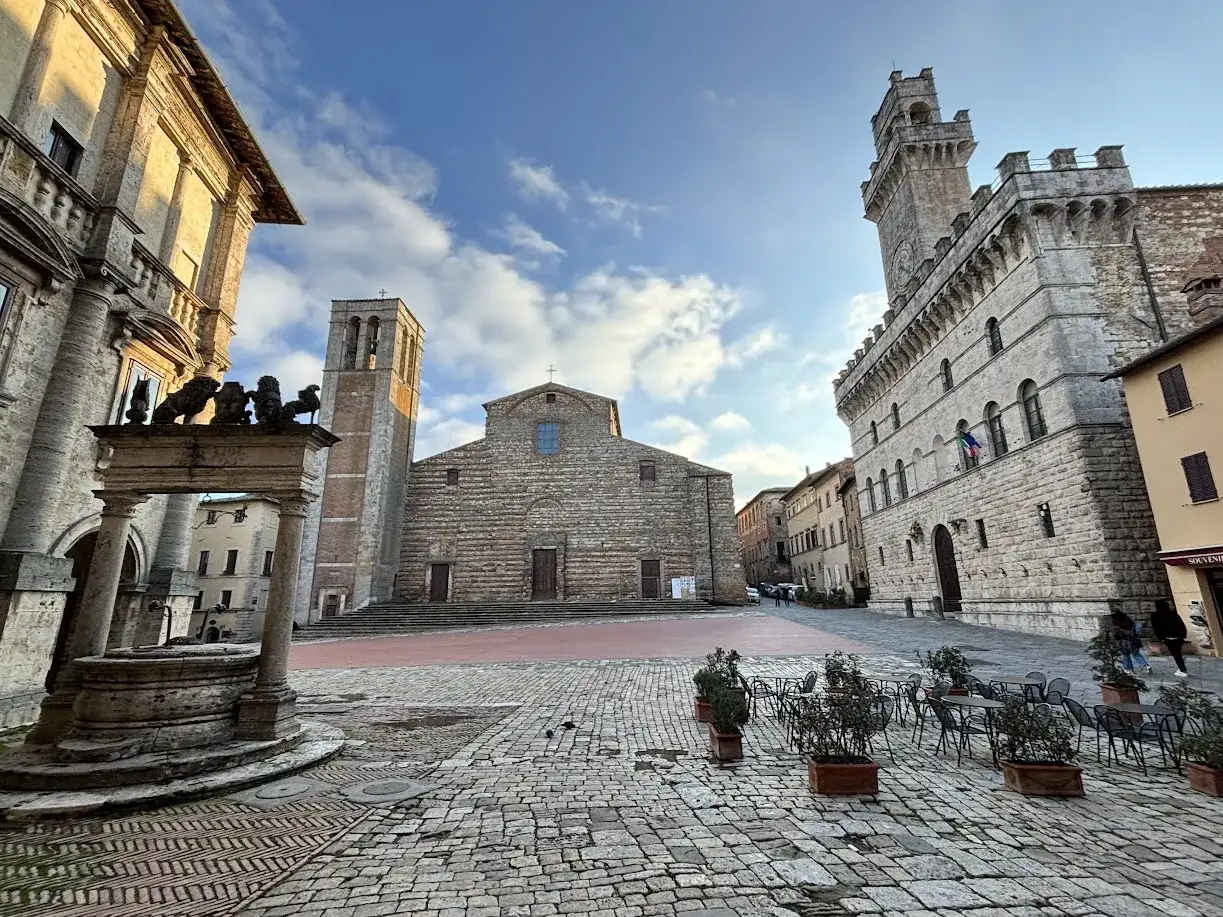 Piazza del Popolo a Montepulciano, con il Duomo, il Palazzo Comunale e il Pozzo dei Grifi sotto un cielo azzurro con nuvole.