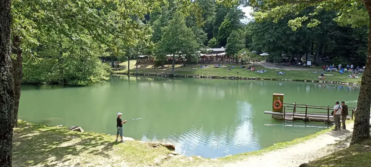 Un ragazzo pesca sul lago Pontini, circondato da alberi e persone che si rilassano sull'erba.