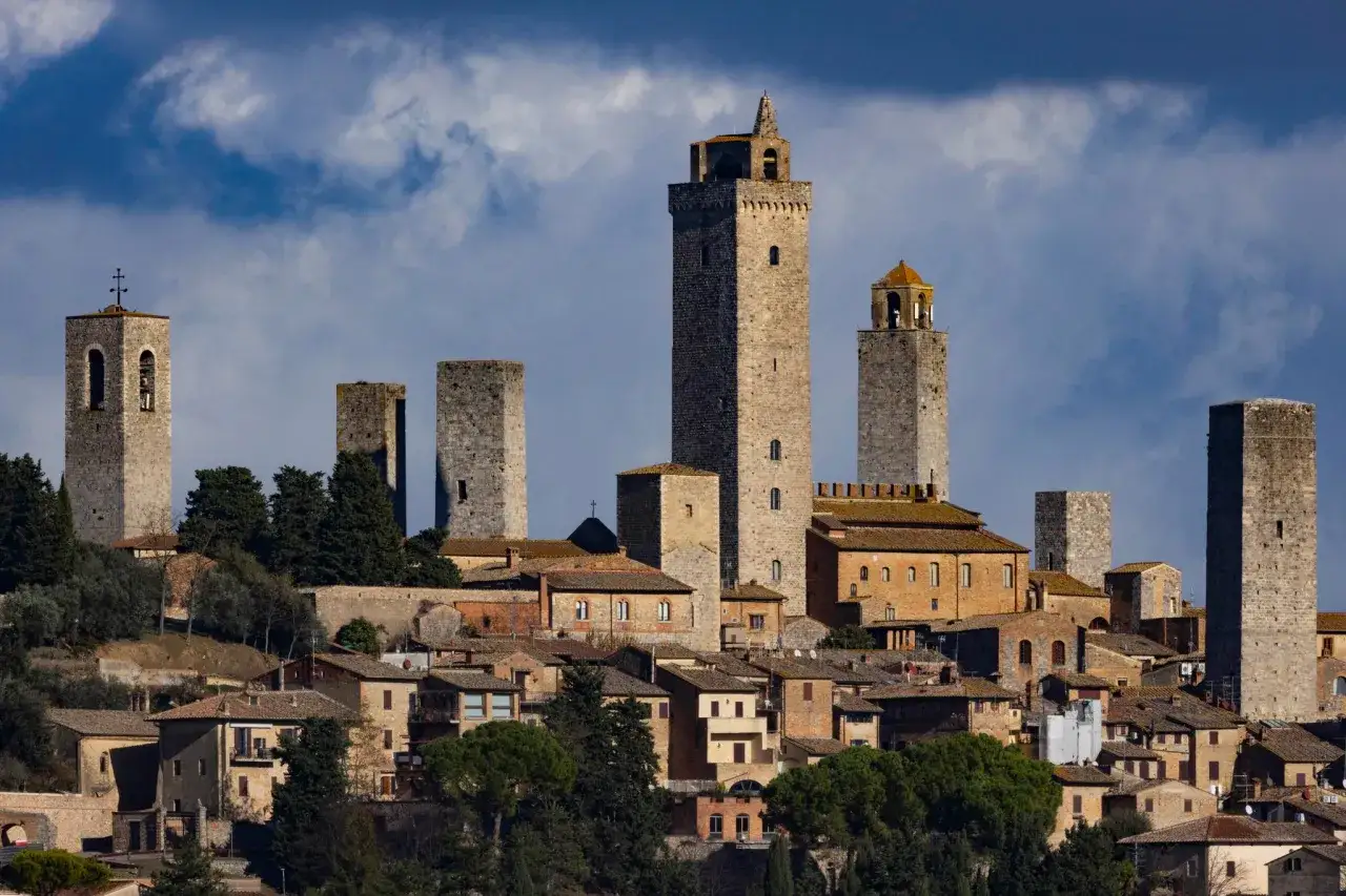 Torri medievali svettano su San Gimignano, un borgo toscano con case in pietra e tetti rossi, sotto un cielo nuvoloso.