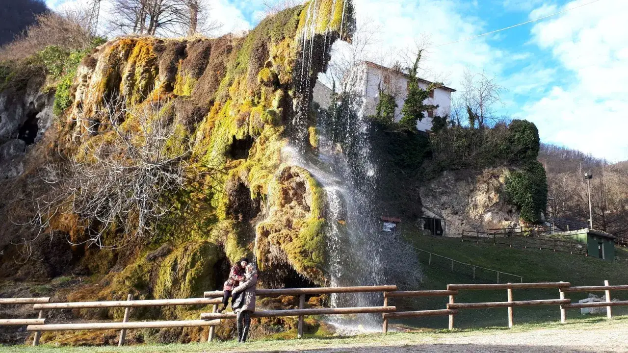 Coppia abbracciata davanti alla cascata delle grotte di Labante, con rocce ricoperte di muschio e un cielo azzurro.