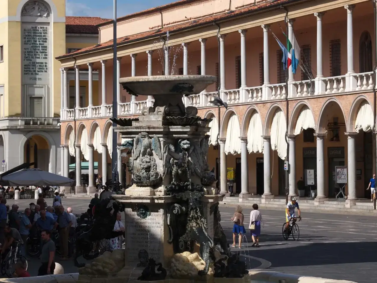 Fontana monumentale in piazza a Faenza, con portici e gente che passeggia.