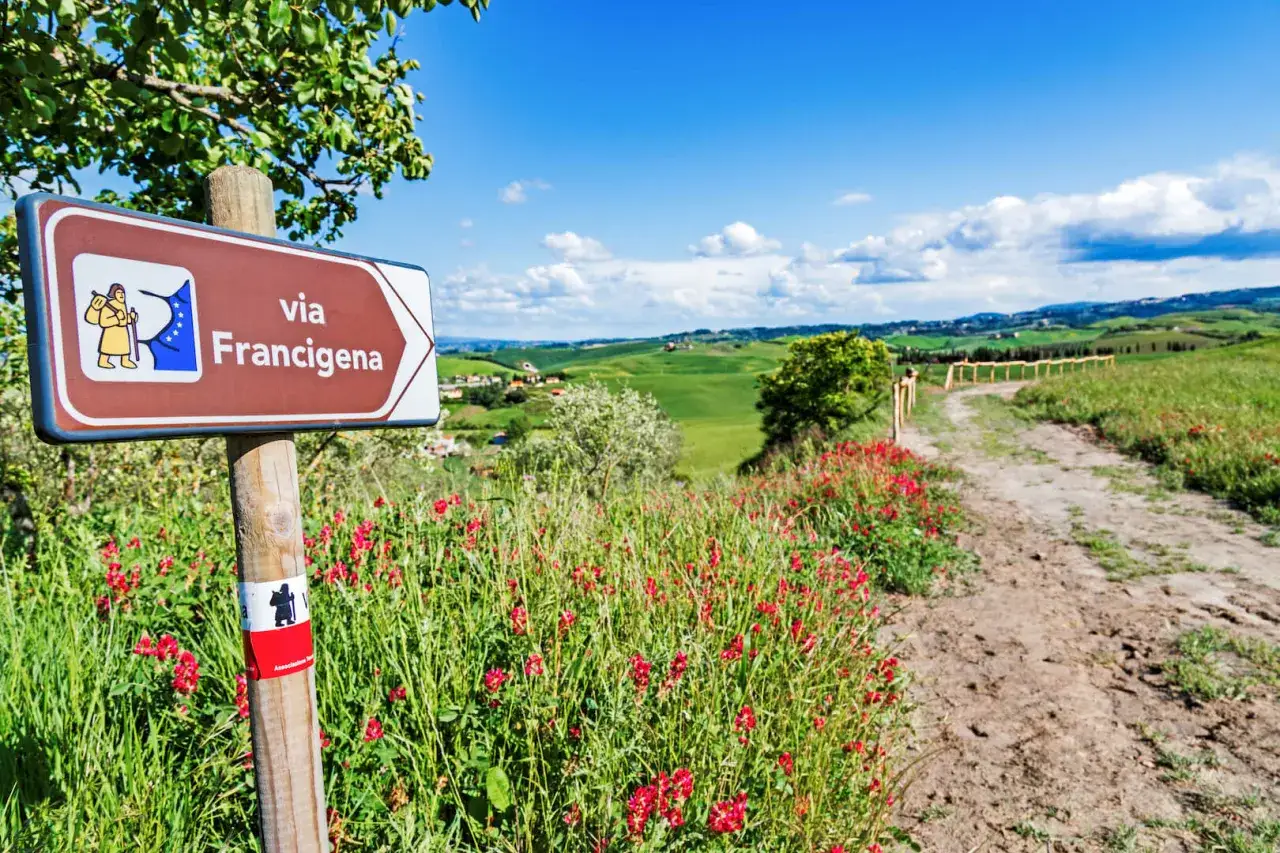 Segnale per il cammino di 3 giorni in Toscana: la Via Francigena attraversa dolci colline verdi e campi fioriti sotto un cielo azzurro.