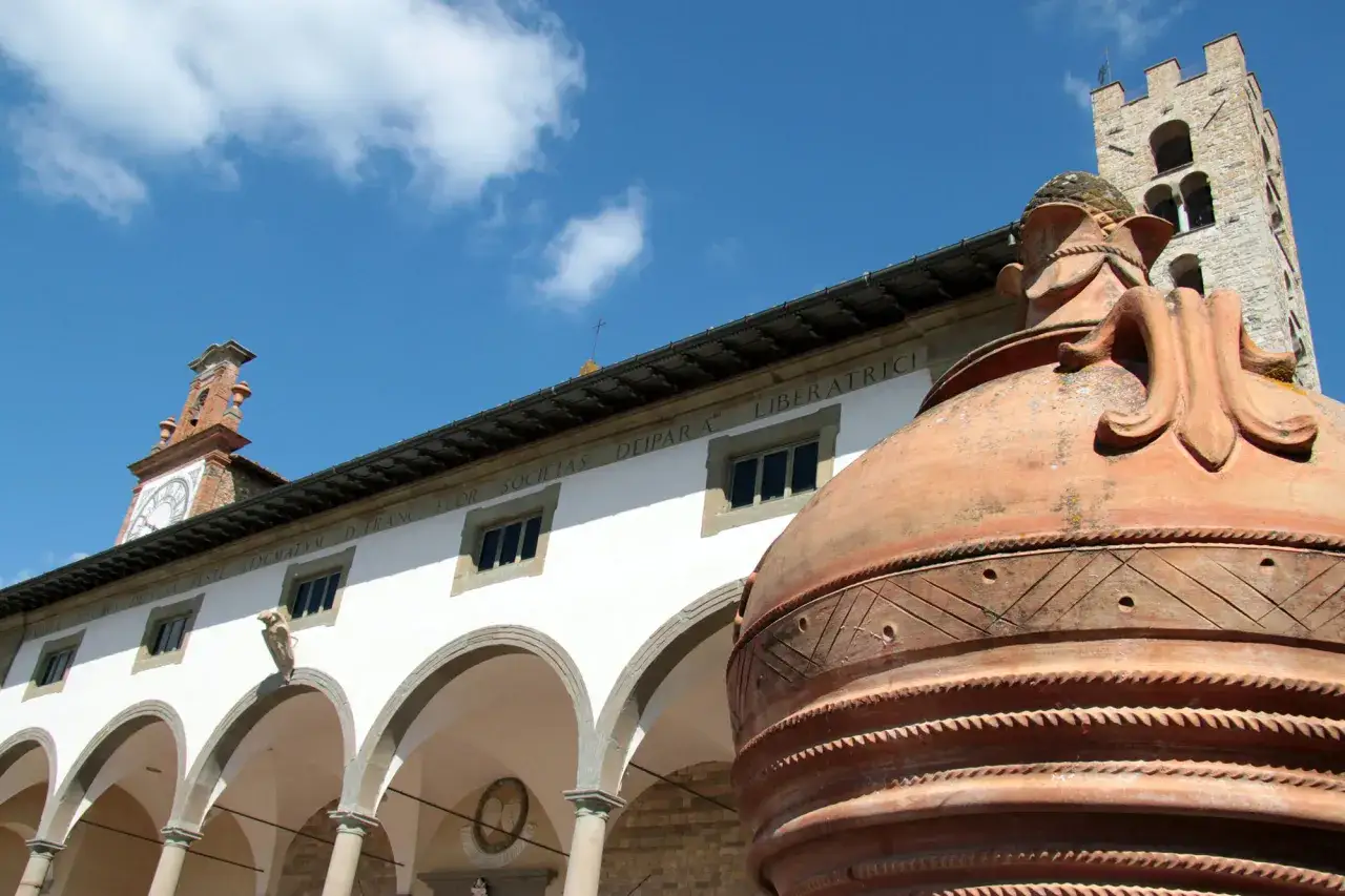 Un'antica anfora in terracotta domina la scena davanti al Palazzo Comunale di Impruneta, Firenze. Un cielo azzurro con nuvole bianche sovrasta la torre dell'orologio.