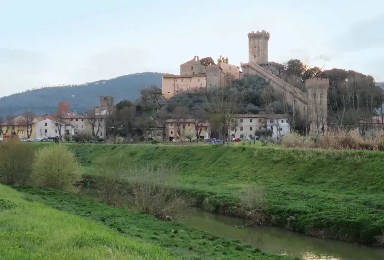Vicopisano cosa vedere: un borgo medievale con rocca e torri domina la collina, con case e un fiume che scorre tra prati verdi.